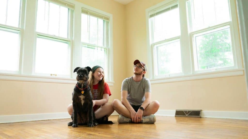 A couple and their dog sitting on a sunlit hardwood floor in an empty room with large windows.