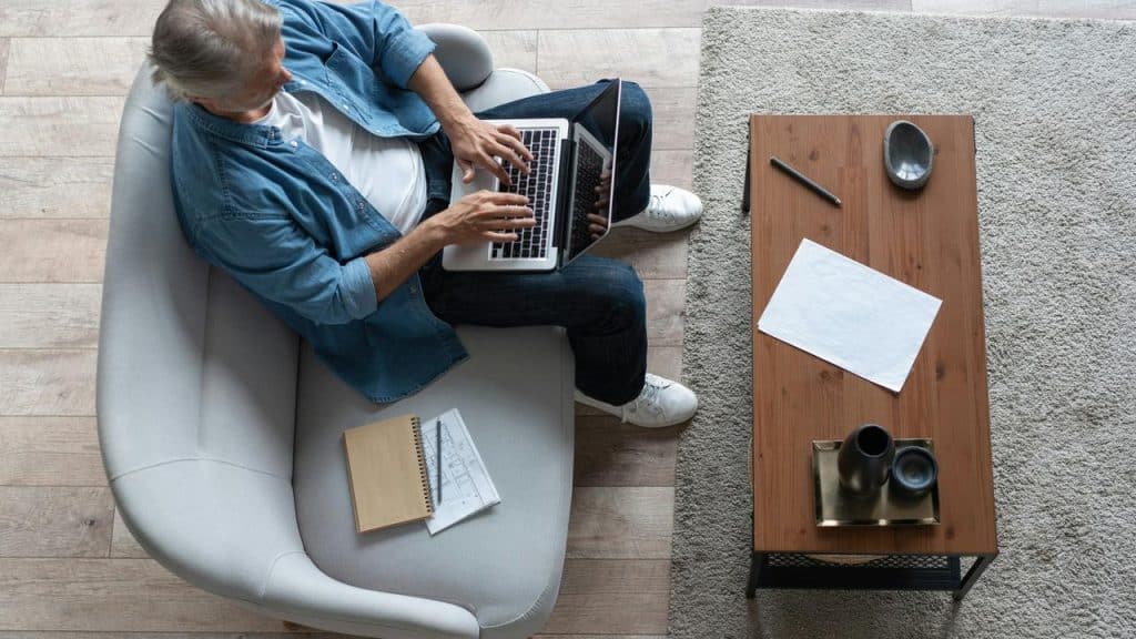 A man on a couch typing on a laptop beside a coffee table.