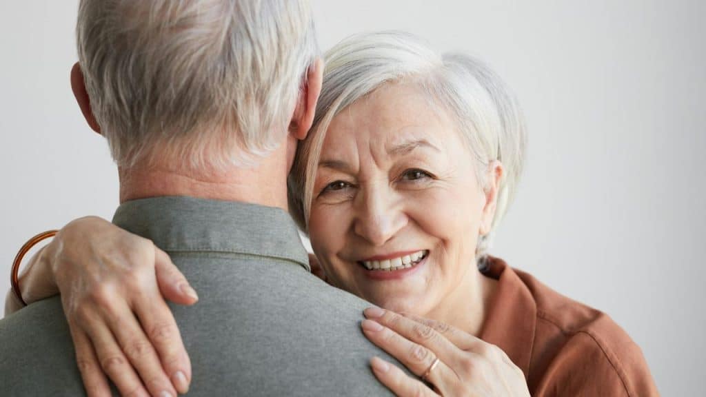 A smiling older woman with white hair embraces a man, looking over his shoulder at the viewer.