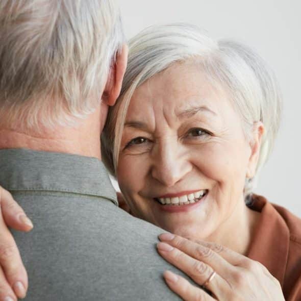 A smiling older woman with white hair embraces a man, looking over his shoulder at the viewer.