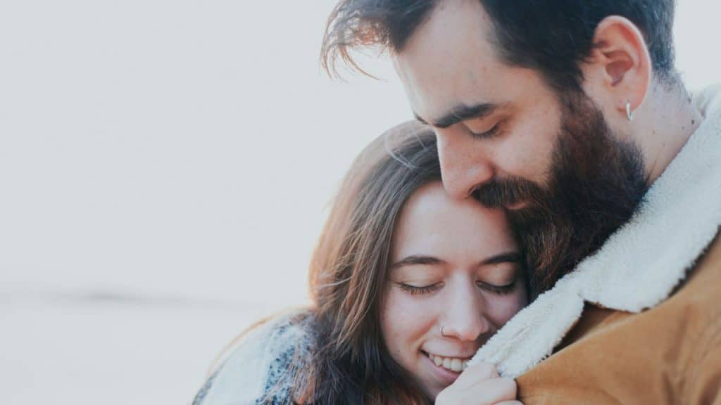 A bearded man embraces a smiling woman, both with their eyes closed, in warm lighting.