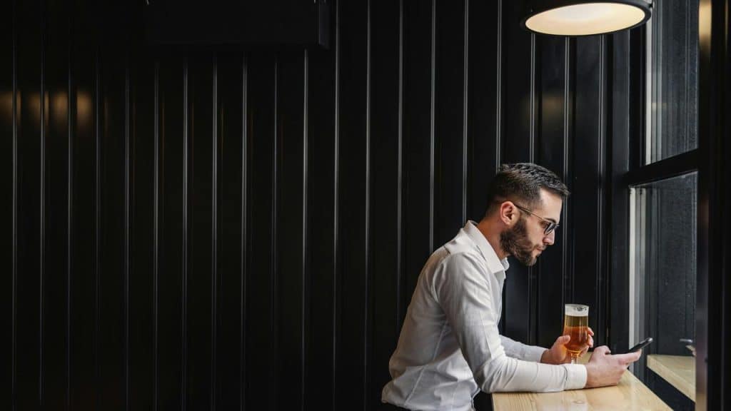 A man holding a beer and looking at his phone by a window.