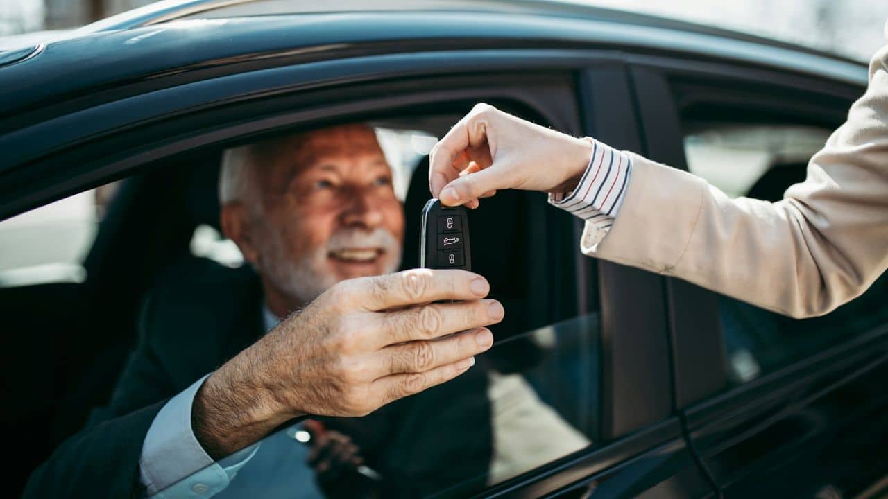 A smiling man in a car receives a car key from another person.