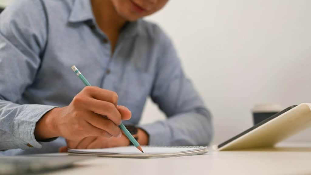 A person in a blue shirt writes in a notebook with a pencil.