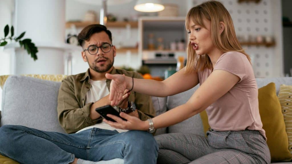 A woman appears to be scolding a man who is looking at his phone while sitting on a sofa.