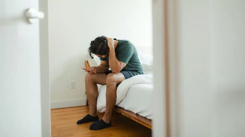 A man sits on the edge of a bed, looking down at his phone with a hand on his head.
