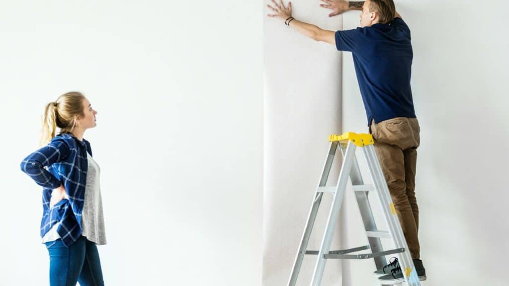 A man on a ladder applies wallpaper while a woman watches him.