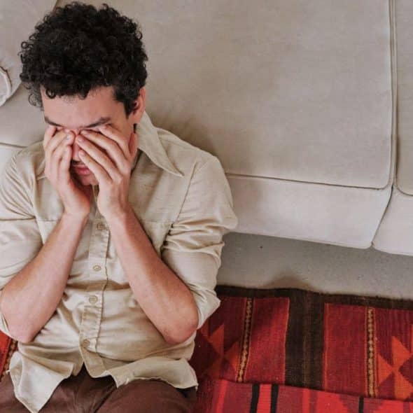 A man with curly hair sits on the floor, rubbing his eyes with both hands.