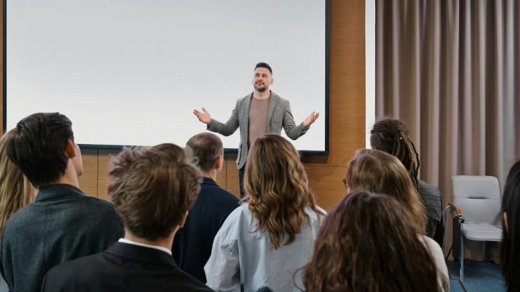 A man with a beard stands on a stage with his arms out, addressing an audience.