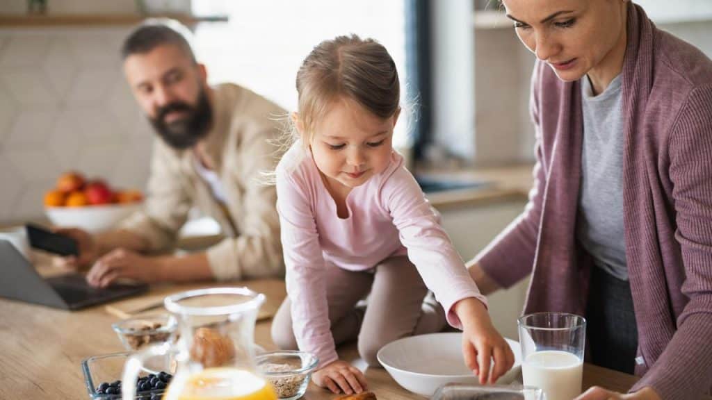 A toddler reaching for breakfast items on the kitchen counter as her parents watch.