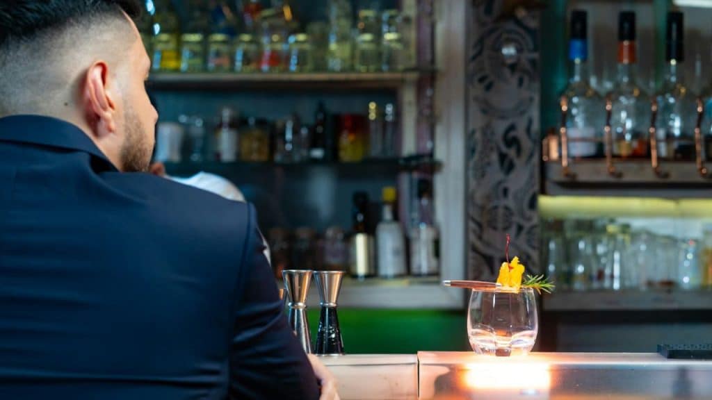 A man in a dark suit sits at a bar counter with a cocktail.