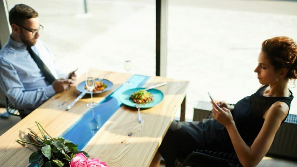 A man and a woman are seated at a table in a restaurant, both looking at their phones.