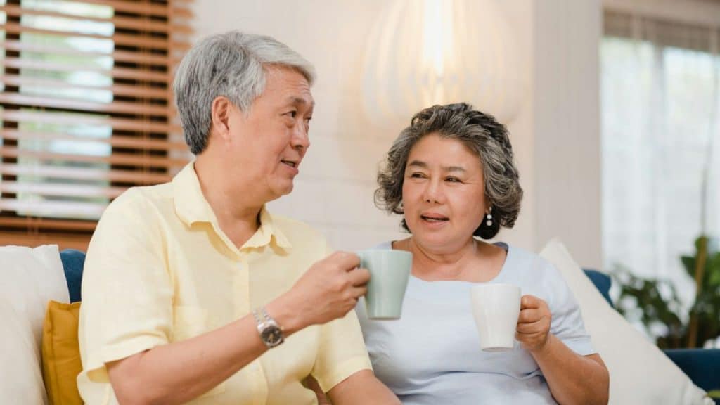 A mature Asian couple happily chats while holding mugs on a sofa.