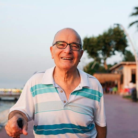 A smiling elderly man with glasses walking along a waterfront promenade.