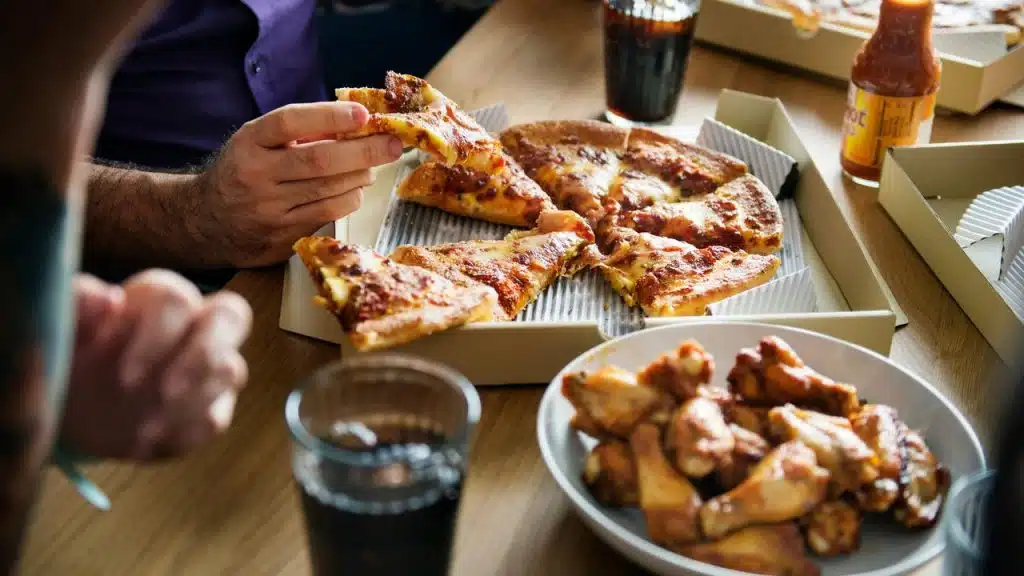 A pepperoni pizza with slices missing, a bowl of chicken wings, and drinks on a table.