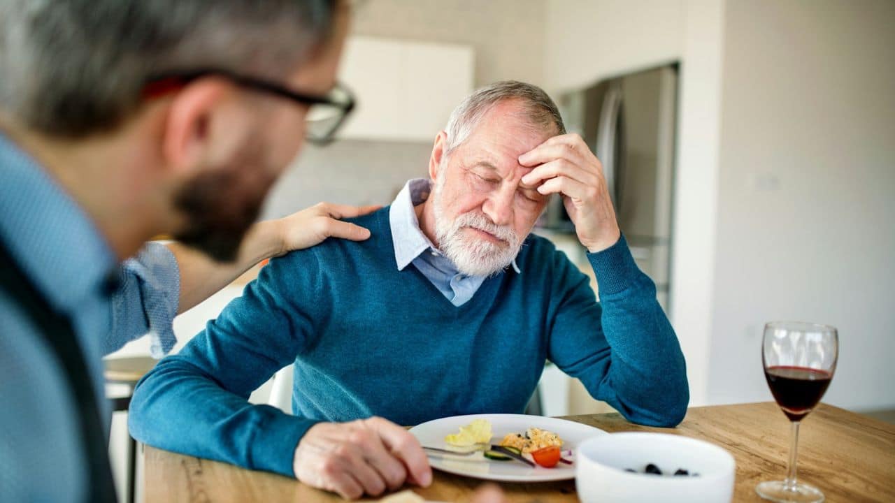 A distressed older man sits at a table while a younger man comforts him.