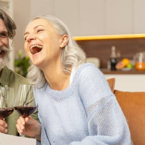 A laughing woman with gray hair holds a wine glass next to a smiling man.