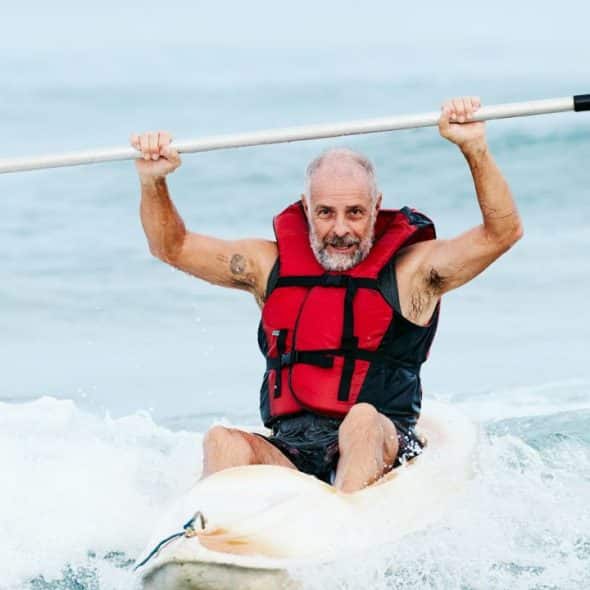 A smiling, bearded man in a red life vest holds a paddle above his head on a kayak.