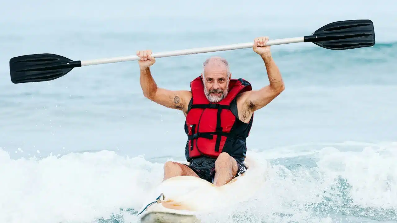 A smiling, bearded man in a red life vest holds a paddle above his head on a kayak.