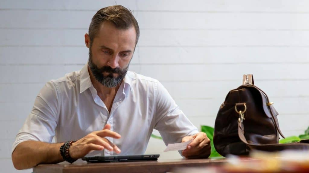 A bearded man in a white shirt looks down at a tablet and a small piece of paper.