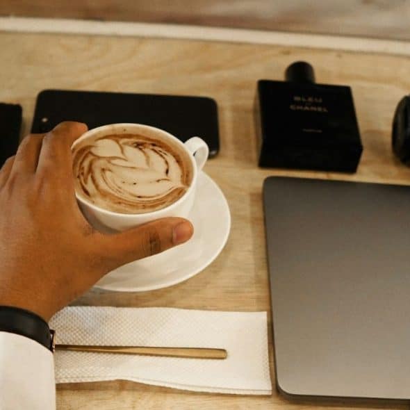 A person's hand reaches for a latte on a wooden table with a laptop and accessories.