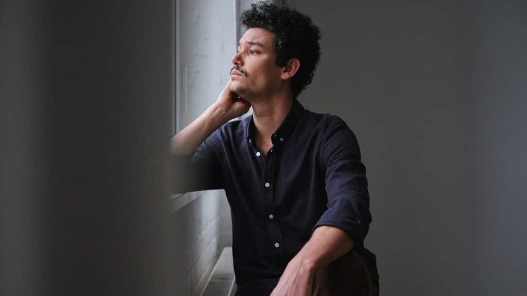 A man with dark, curly hair and a mustache looks out a window.