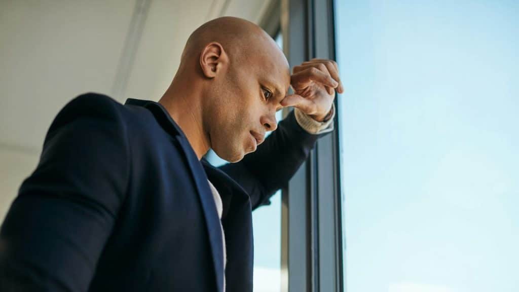 A bald man in a suit jacket leans against a window, looking downcast.