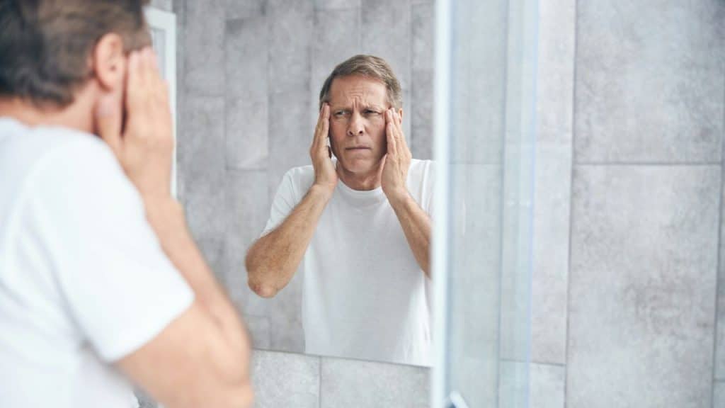 A distressed man in a white t-shirt looks into a mirror, holding his temples.