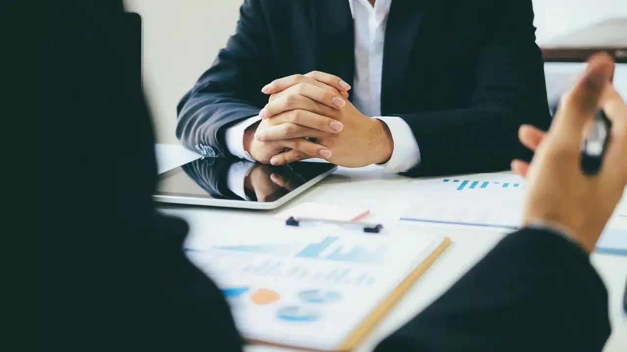 A suited person with clasped hands over a tablet and charts on a desk.