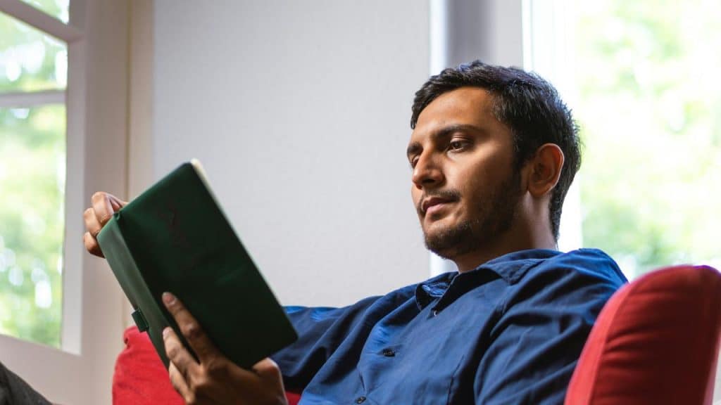 A man seated on a red couch reading a green hardcover book by a window.
