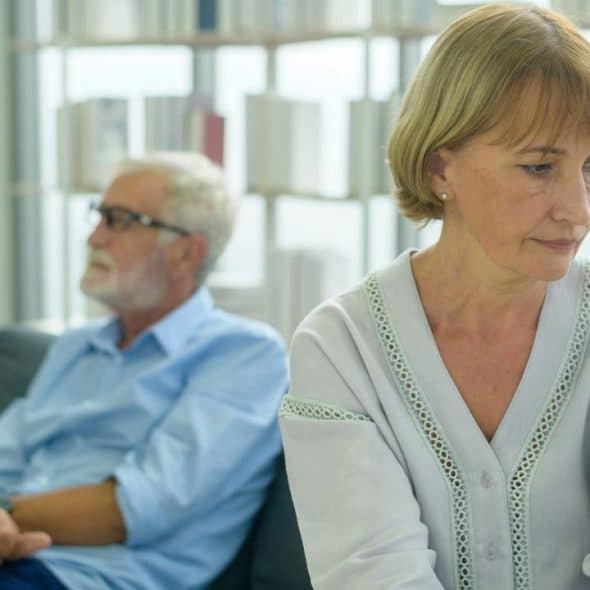 A somber woman looks down while a man sits on a couch behind her, turned away.