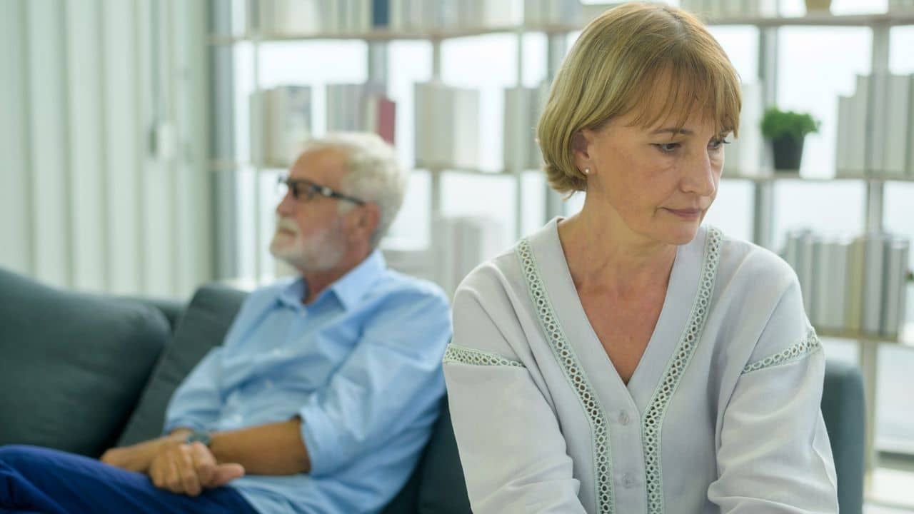 A somber woman looks down while a man sits on a couch behind her, turned away.