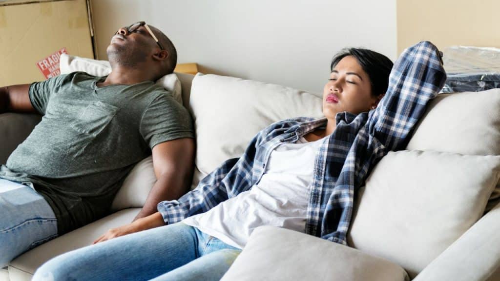 A man and woman are asleep on a white couch amidst moving boxes.