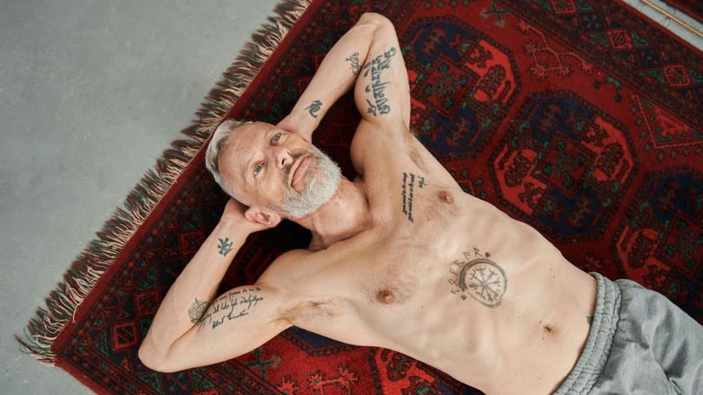 A man with a white beard and tattoos on his arms and chest lies on a red patterned rug.