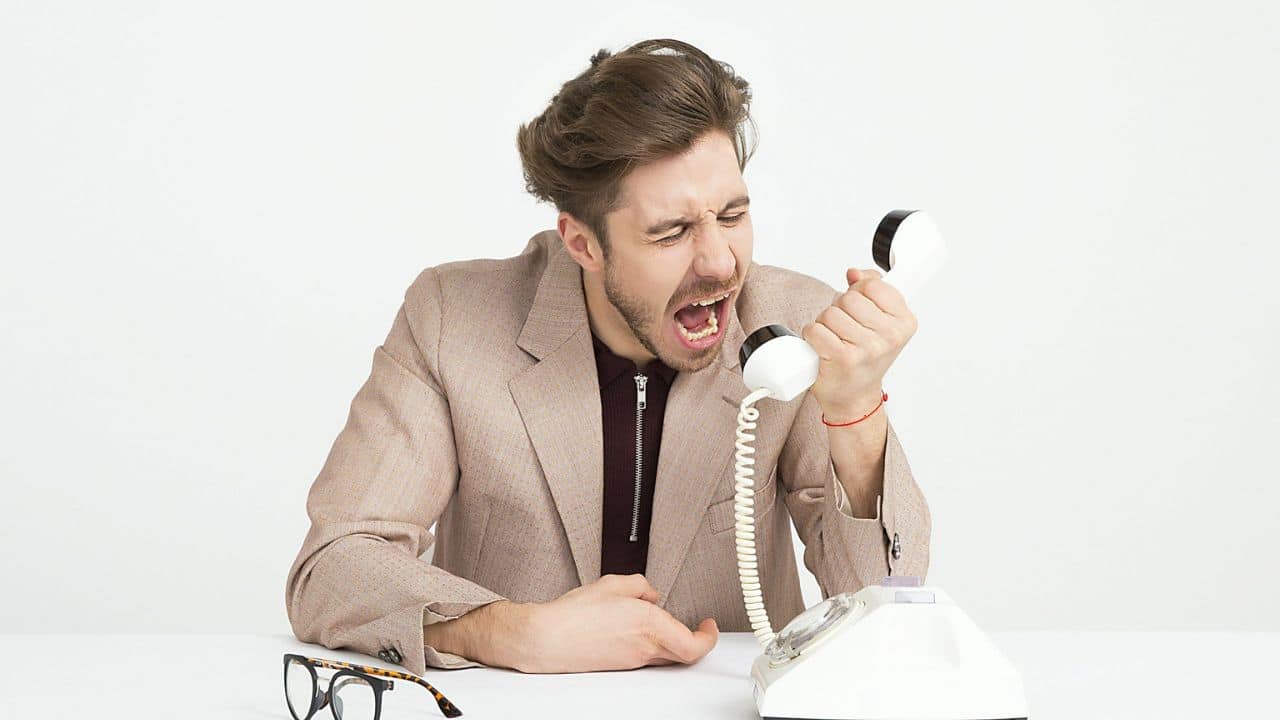 A man in a suit screams into a white landline phone, with glasses on the table.