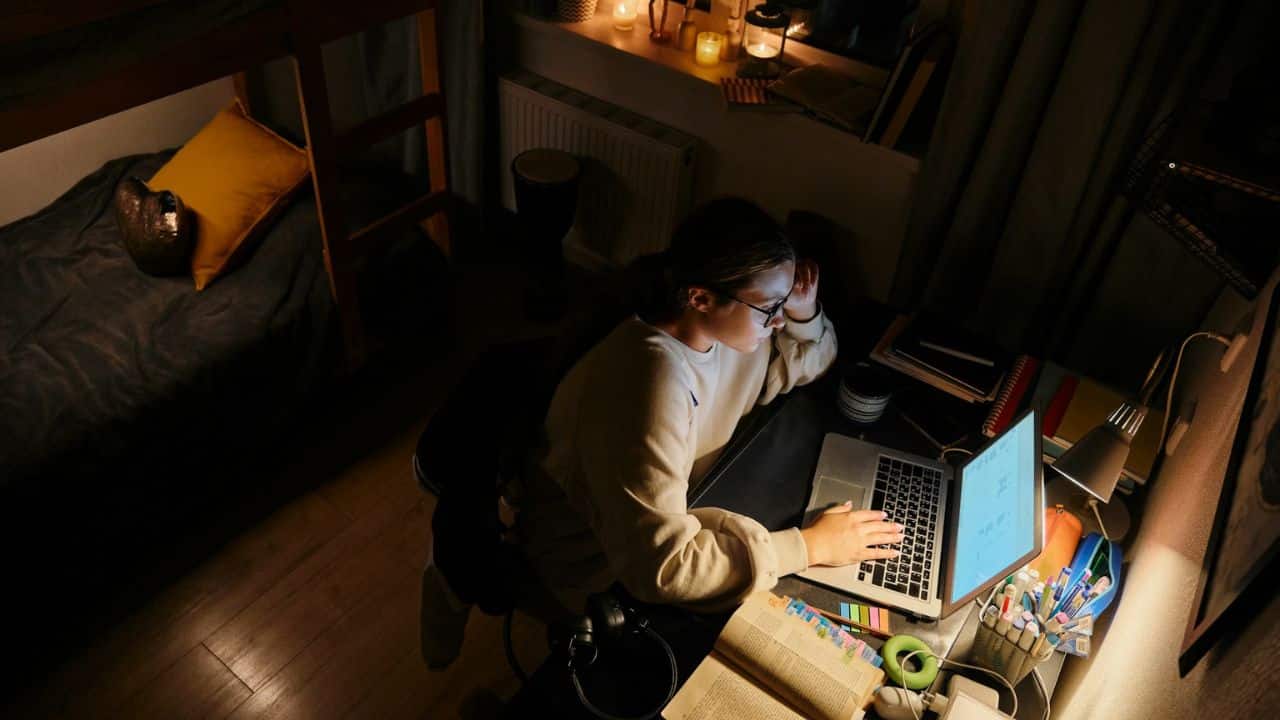 A person wearing glasses works on a laptop at a desk in a dimly lit room with a bed nearby.