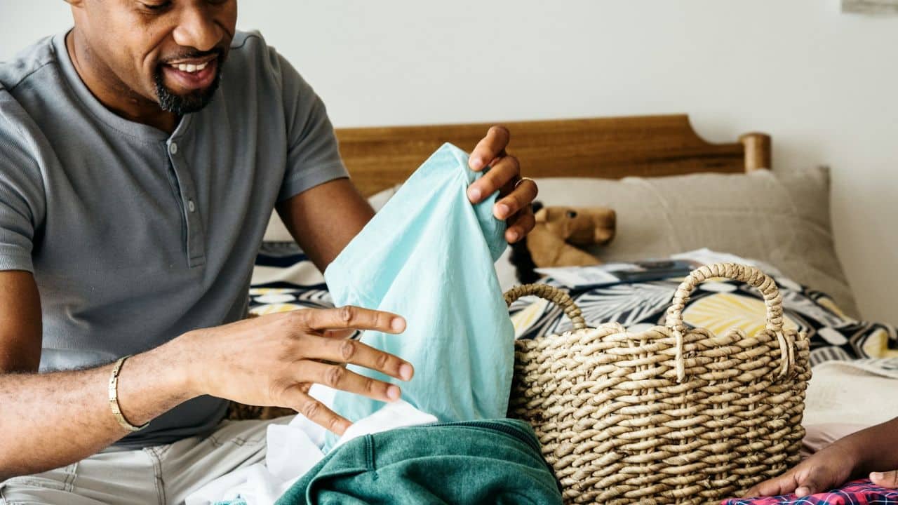 A smiling man folds light blue laundry, next to a wicker basket on a bed.