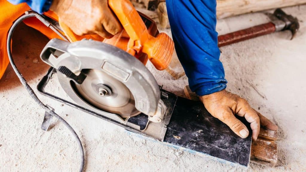 A worker’s hand guiding an orange circular saw as it cuts a plank on a dusty floor.