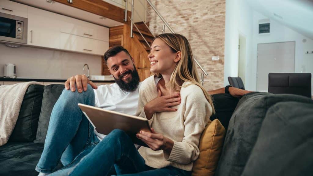 A couple on a sofa laughing together while looking at a tablet in their living room.