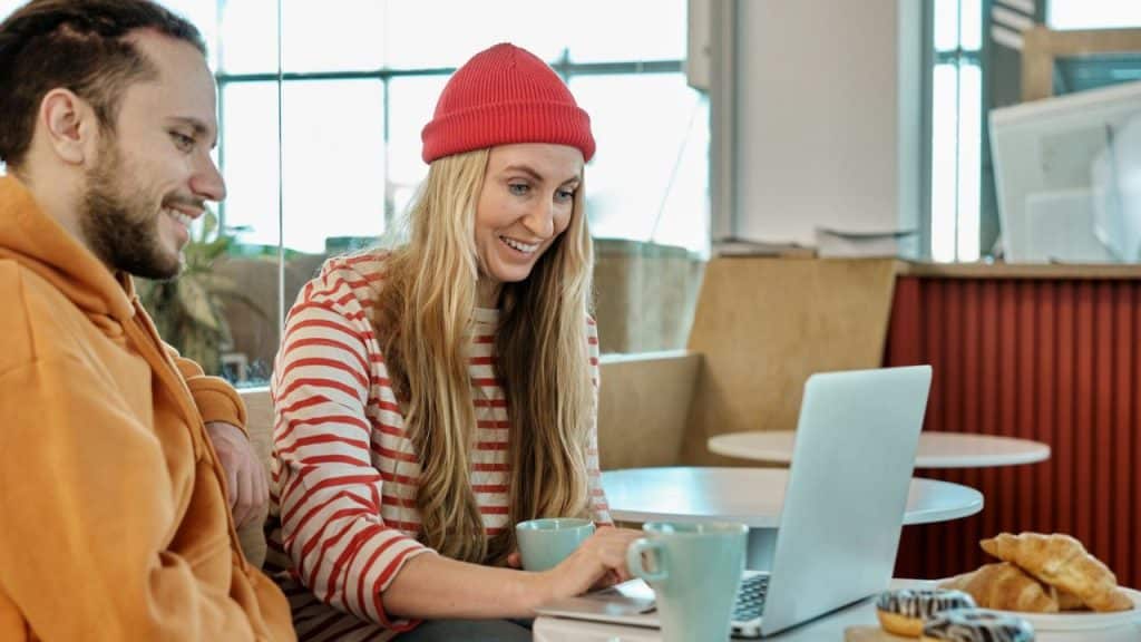 A man and woman smiling at a laptop, with pastries and coffee mugs on the table.