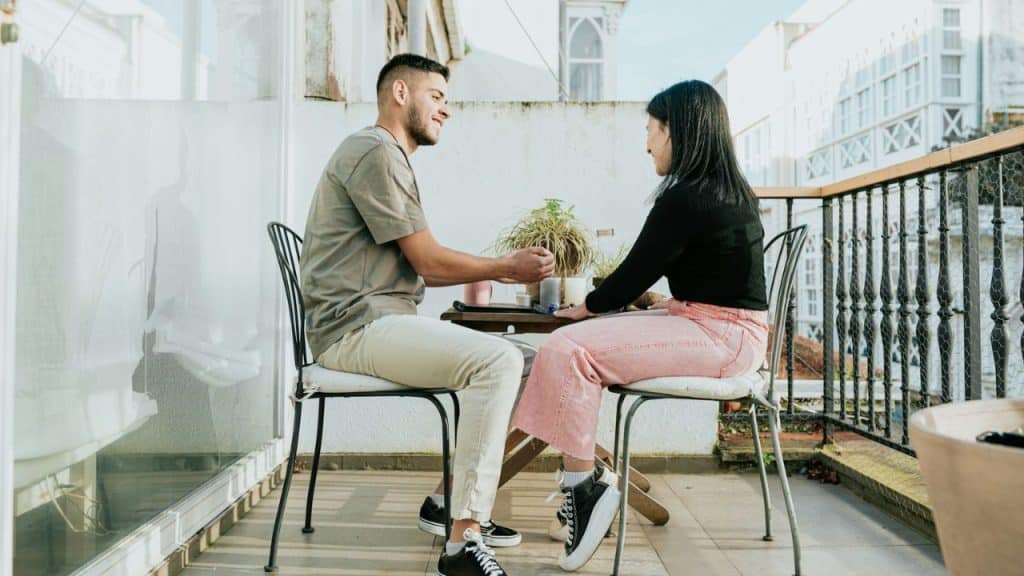 A man and a woman sit at a small table on a balcony, looking at each other and smiling.