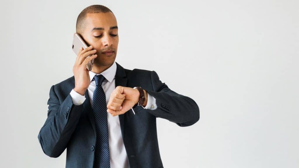 A man in a suit talks on a phone while checking his watch.