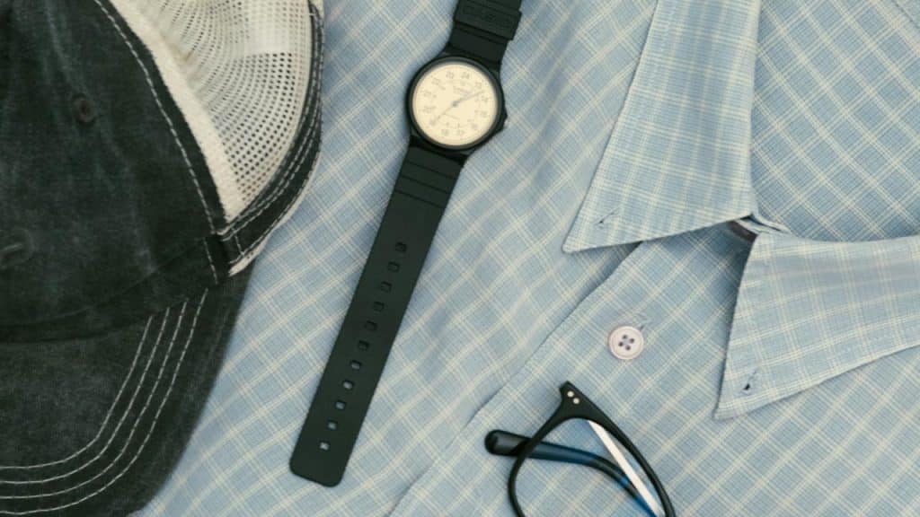 A black wristwatch on a blue checkered shirt beside a cap and glasses.