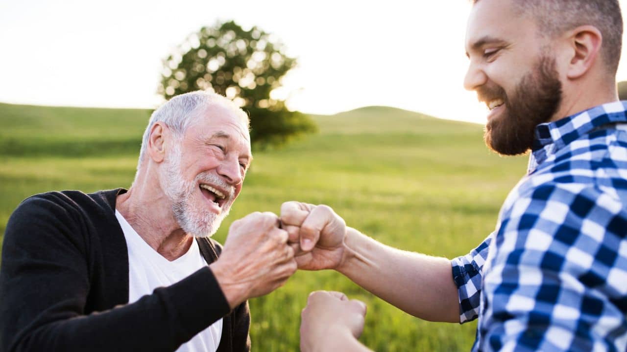A laughing older man fist bumps a younger, bearded man outdoors.