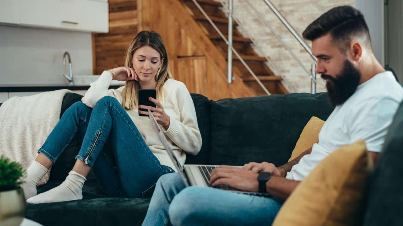 A couple on a couch, a woman on her smartphone and a man on his laptop.