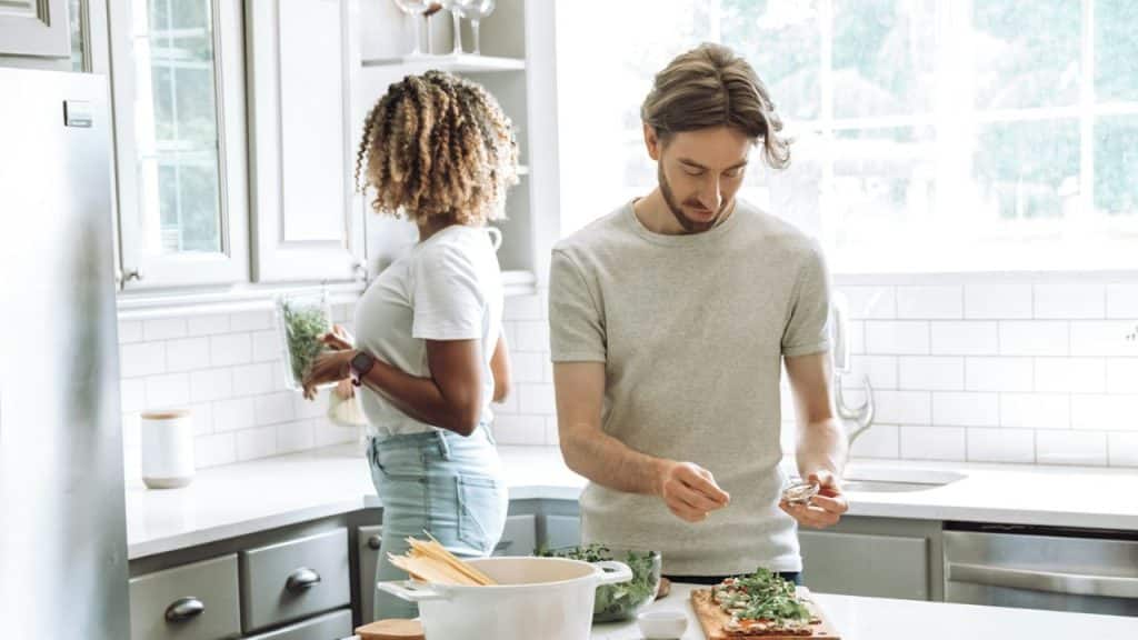 A couple in a bright kitchen prepares food; the man is adding ingredients, and the woman is by the refrigerator.