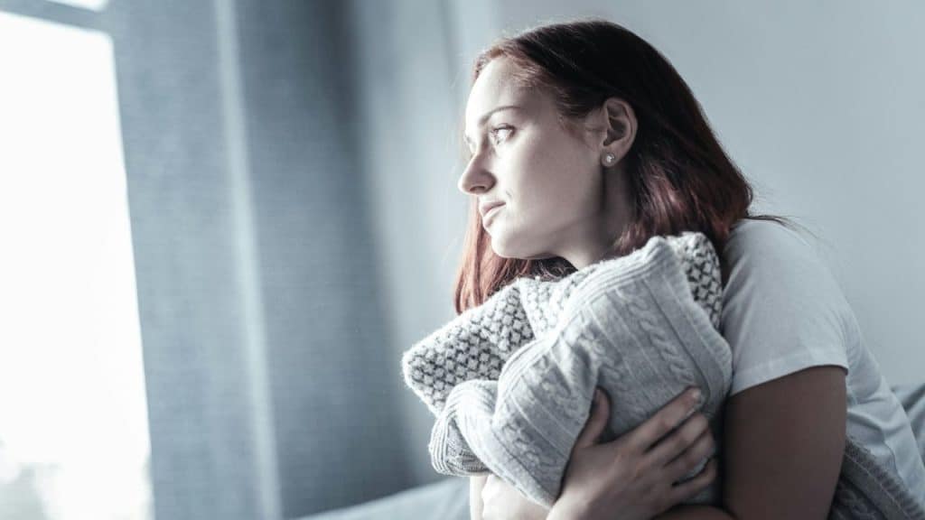 A pensive woman with reddish hair holds a knitted pillow, looking out a window.