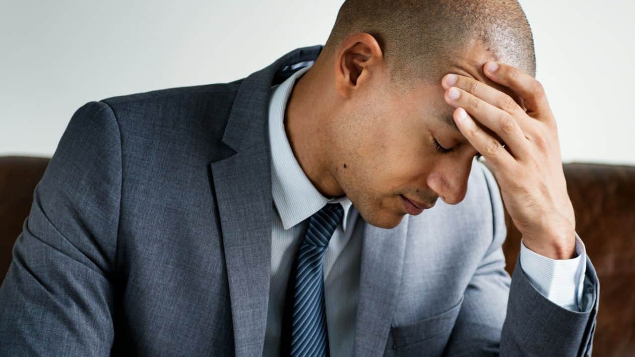 A man in a suit and tie sits with his hand on his forehead, looking downcast.