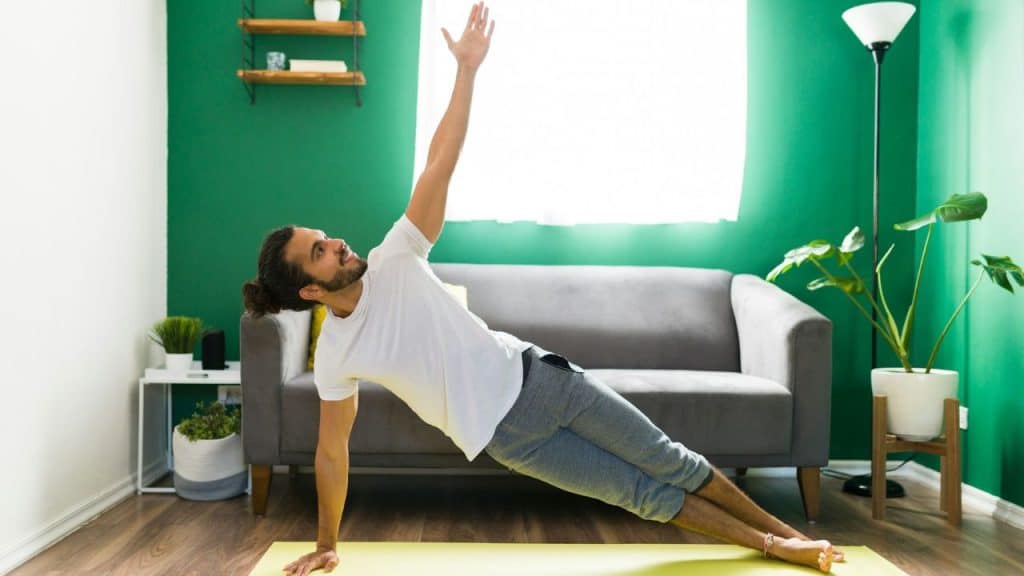 A man with a beard and long hair holds a side plank pose on a yoga mat.