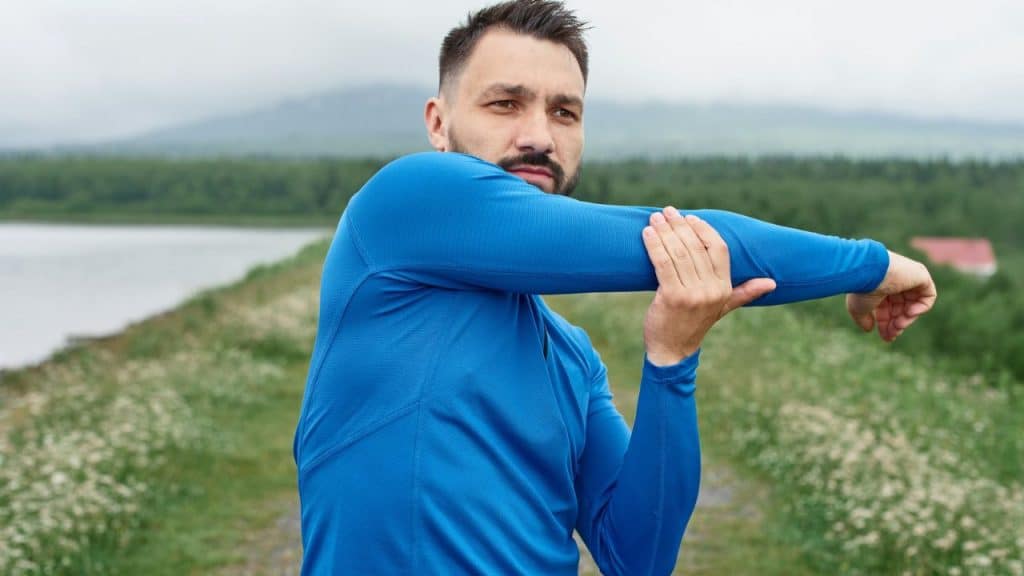 A man in a blue long-sleeved shirt stretches his arm across his body outdoors.